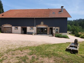 Ein zweistöckiges Gebäude mit rotem Ziegeldach, einem Hauseingang mit Stufen, einer Bank vor der Fassade und einem kleinen Gartenbereich mit Wasserspiel.