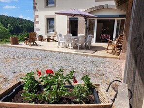 Eine Holzterrasse mit Gartenmöbeln, ein erhöhter Blumenkasten mit Pflanzen und ein Gebäude mit einer Steinaußenfassade.