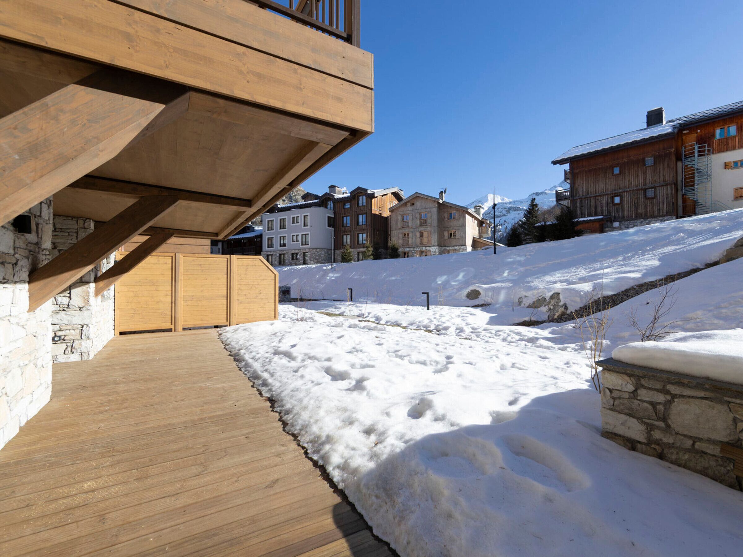A wooden deck with a stone wall, snow-covered ground, and buildings in the background.