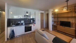 Eine moderne Küche mit weißen Schränken, einem eingebauten Backofen und einer Kaffeemaschine. Im Wohnbereich befindet sich ein Holzessttisch und eine Couch, auf der ein Holzregal mit einem Fernseher steht.
