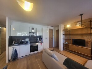 Eine moderne Küche mit weißen Schränken, einem eingebauten Backofen und einer Kaffeemaschine. Im Wohnbereich befindet sich ein Holzessttisch und eine Couch, auf der ein Holzregal mit einem Fernseher steht.
