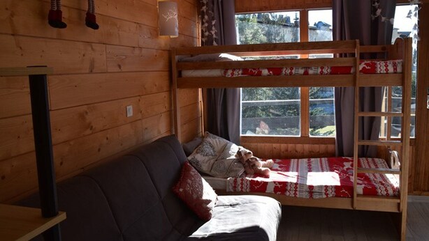 Ein Etagenbett mit einer rot-weißen Bettdecke, eine Holzwand, ein Sofa mit einem roten Kissen und ein Fenster mit Vorhängen.