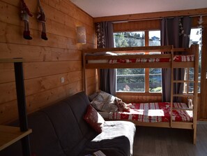 A bunk bed with a red and white patterned bedspread, a wooden wall, a sofa with a red pillow, and a window with curtains. - Central studio in Montgenèvre - 2 guests (Montgenèvre)