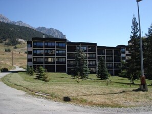 A multi-story building with a modern architectural design, surrounded by a grassy area with a few trees and a mountain range in the background. - Central studio in Montgenèvre - 2 guests (Montgenèvre)