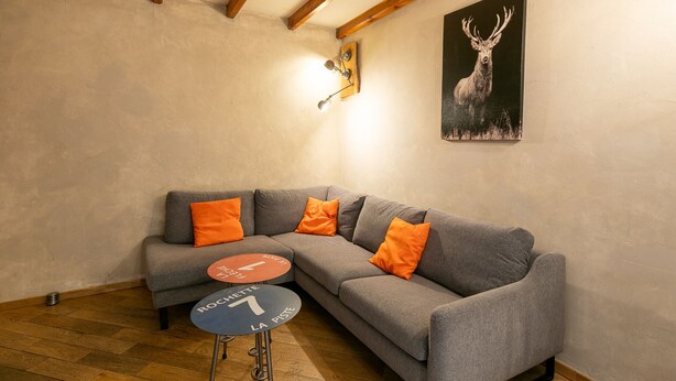 Una habitación con un sofá gris, cojines naranjas, una pequeña mesa redonda, una lámpara de pared y un cuadro enmarcado de un ciervo.