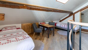 Un dormitorio tipo buhardilla acogedor, con techo inclinado, vigas de madera, una pequeña mesa de comedor, dos sillas, una cama con ropa de cama estampada y un tragaluz.