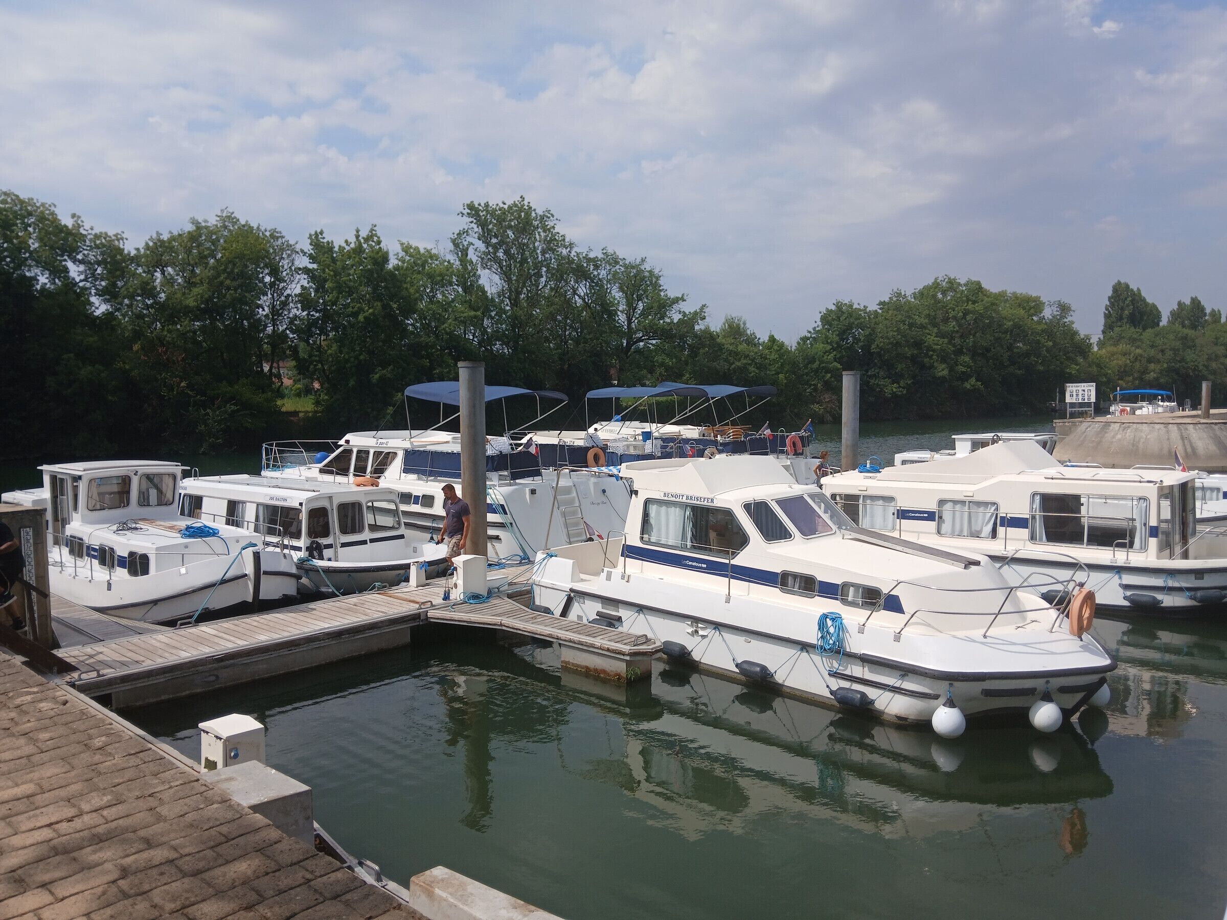 A marina with several boats docked, a wooden dock, and a calm water body surrounded by trees.