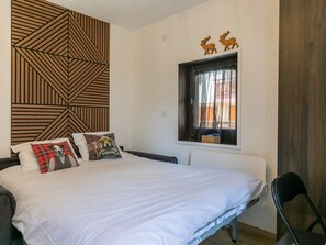 A bedroom with a bed, pillows, a window with curtains, a wooden wall panel, and a small wooden shelf. - Cozy studio in the heart of the station (Le Monêtier-les-Bains)