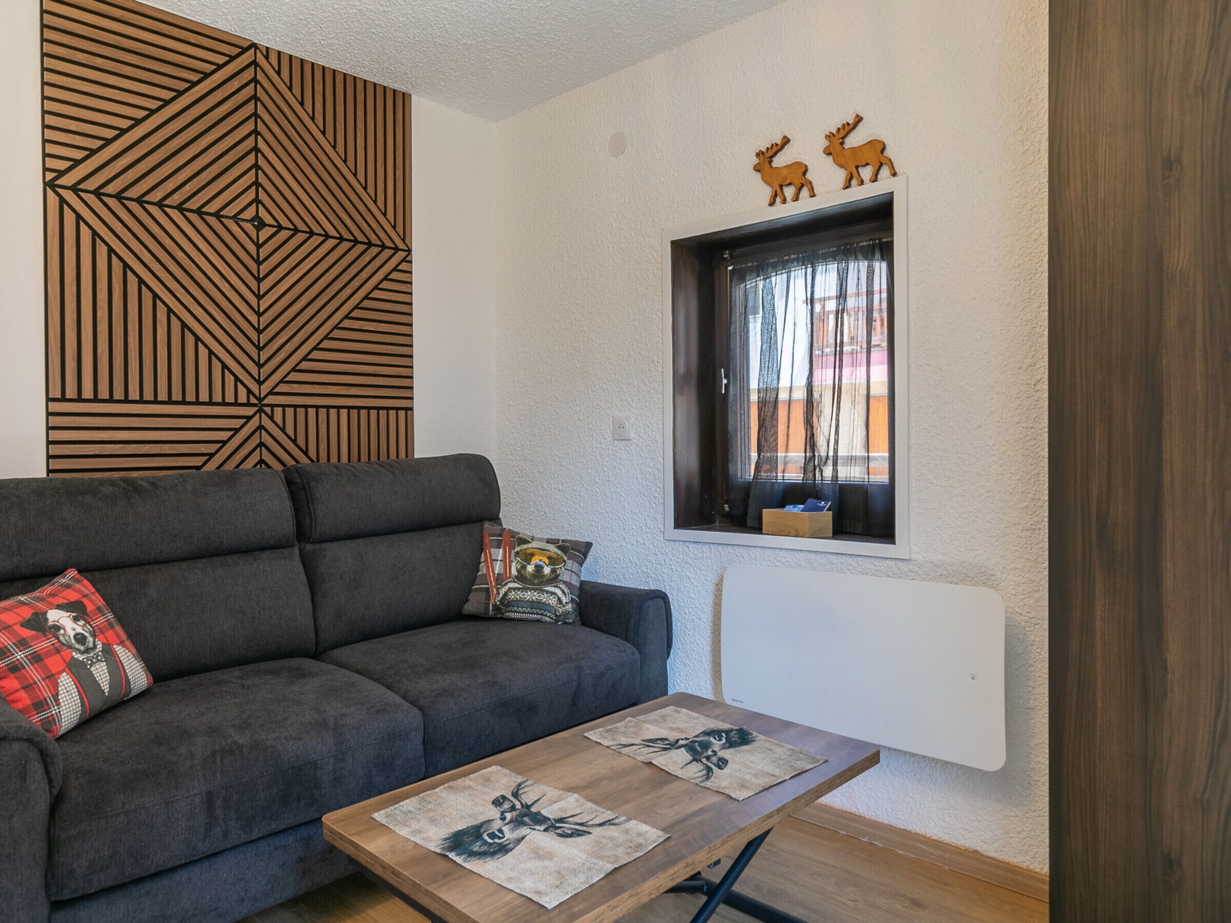 Een moderne woonkamer met een bank, een bijzettafeltje met dierenprint, een geometrisch wandpaneel, een raam met gordijnen en een wandplank met decoratieve objecten.