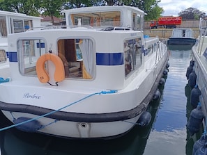 Un barco llamado 'Perdrix' atracado en un puerto deportivo, con un flotador de salvamento sujeto a la popa, ventanas y una cabina con cortinas.