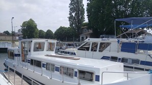 Un bateau blanc était amarré dans une marina. Il était doté d’une terrasse couverte, de fenêtres sur les côtés et d’une structure de cabine bien visible.