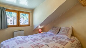 Ett sovrum med ett sluttande tak, ett fönster med gardiner, ett element, ett nattduksbord med en lampa och en säng med mönstrat sänglinne.