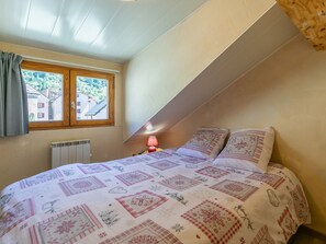 Ein Schlafzimmer mit geneigter Decke, einem Fenster mit Vorhängen, einem Heizkörper, einem Nachttisch mit Lampe und einem Bett mit gemusterten Betttextilien.