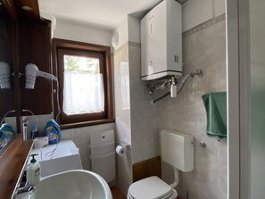 Casa de banho compacta com sanita, lavatório e chuveiro. Há uma janela que proporciona luz natural e uma toalha pendurada na parede.