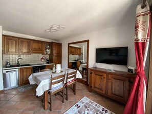 Cozinha compacta com armários de madeira, mesa de jantar com cadeiras, forno embutido e televisão de tela plana presa na parede.