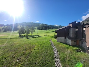 A modern building with a stone base and dark upper structure, surrounded by a green field and trees. - 3-bedroom apartment on the slopes - 6 guests (Montgenèvre)