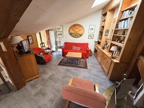A cozy living room with a red sofa, a wooden coffee table, and a checkered chair. A bookshelf filled with books and decorative items is to the right. - 3-bedroom apartment on the slopes - 6 guests (Montgenèvre)