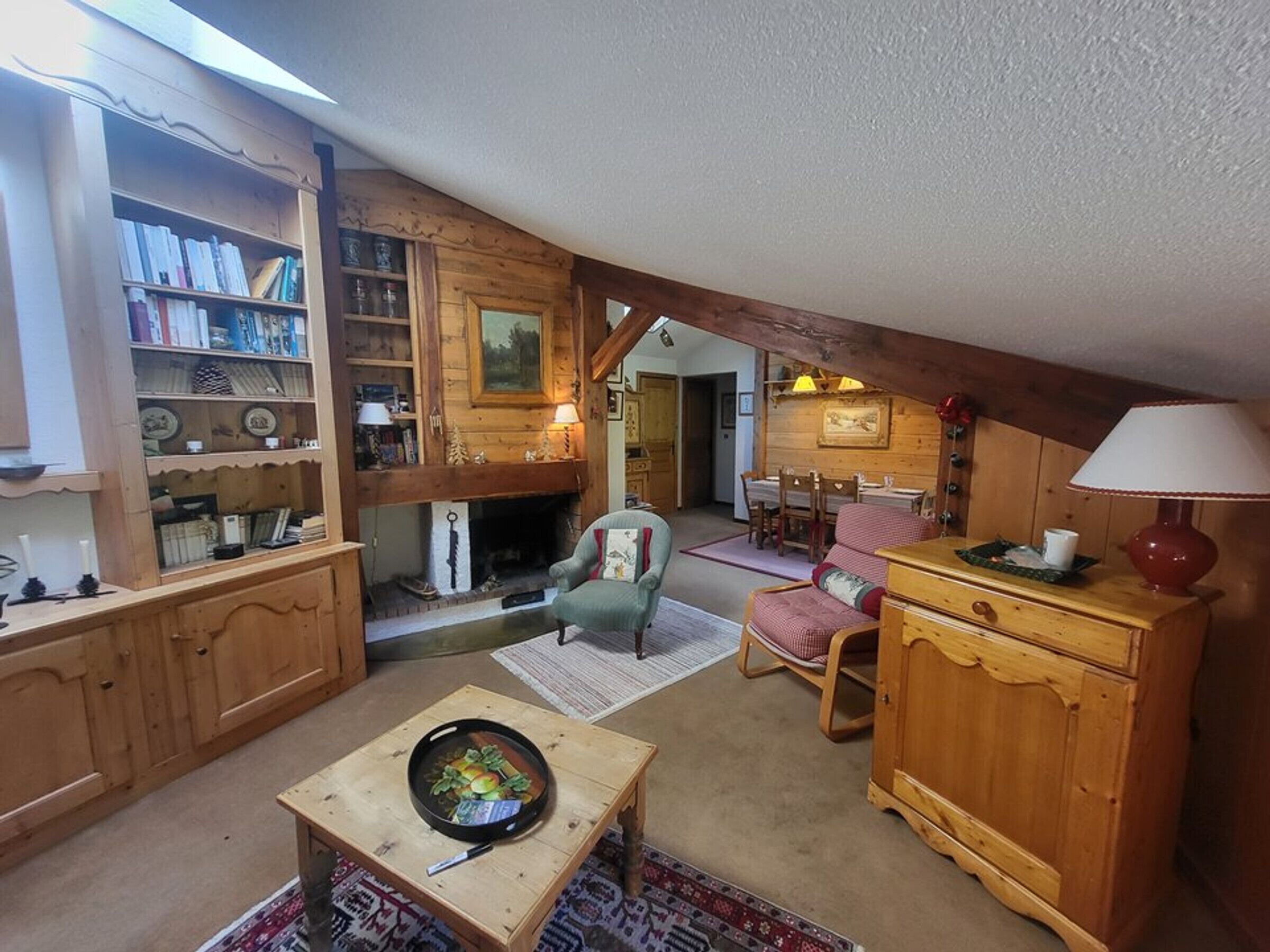Un salon chaleureux, aménagé avec du mobilier en bois, doté d’une cheminée, d’un coin repas et d’une bibliothèque remplie de livres et d’objets décoratifs.