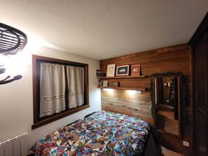 Ein Schlafzimmer mit Holz-Kopfteil, einem Bett mit farbenfroher Steudecke, einem Fenster mit Vorhängen und einem Regal mit gerahmten Bildern.