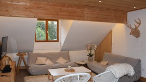 Un salon moderne avec un plafond en bois, un canapé gris, des chaises blanches, une petite table et une fenêtre donnant sur des arbres.
