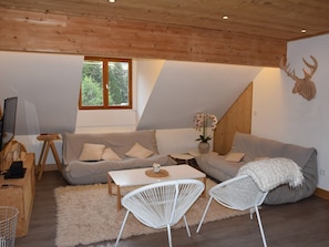 Ein modernes Wohnzimmer mit Holzboden, einem grauen Sofa, weißen Stühlen, einem kleinen Tisch und einem Fenster mit Blick auf Bäume.