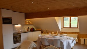 Un coin repas chaleureux avec une table dressée pour quatre personnes, une cuisine équipée d’appareils modernes et une grande fenêtre laissant entrer la lumière naturelle.