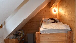 Une chambre mansardée chaleureuse avec un plafond en pente, des murs en bois, un lit simple avec une couette blanche et une petite étagère avec des livres et un panier.