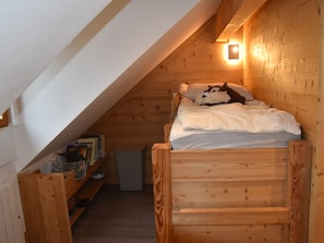 Ein gemütliches Schlafzimmer im Dachgeschoss mit schrägen Wänden, Holzwänden, einem Einzelbett mit weißer Decke und einem kleinen Regal mit Büchern und einem Korb.
