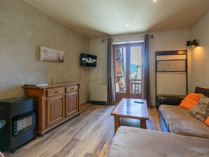 A room with a wooden cabinet, a television, a sofa with orange cushions, a table, and a view through a door. - Cozy duplex in village center (Le Monêtier-les-Bains)