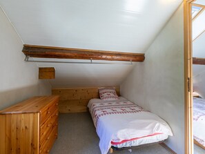 Ein kleines Schlafzimmer mit geneigtem Dach, einem hölzernen Schminktisch, einem Bett mit gemusterten Betttextilien und einem Dachfenster.