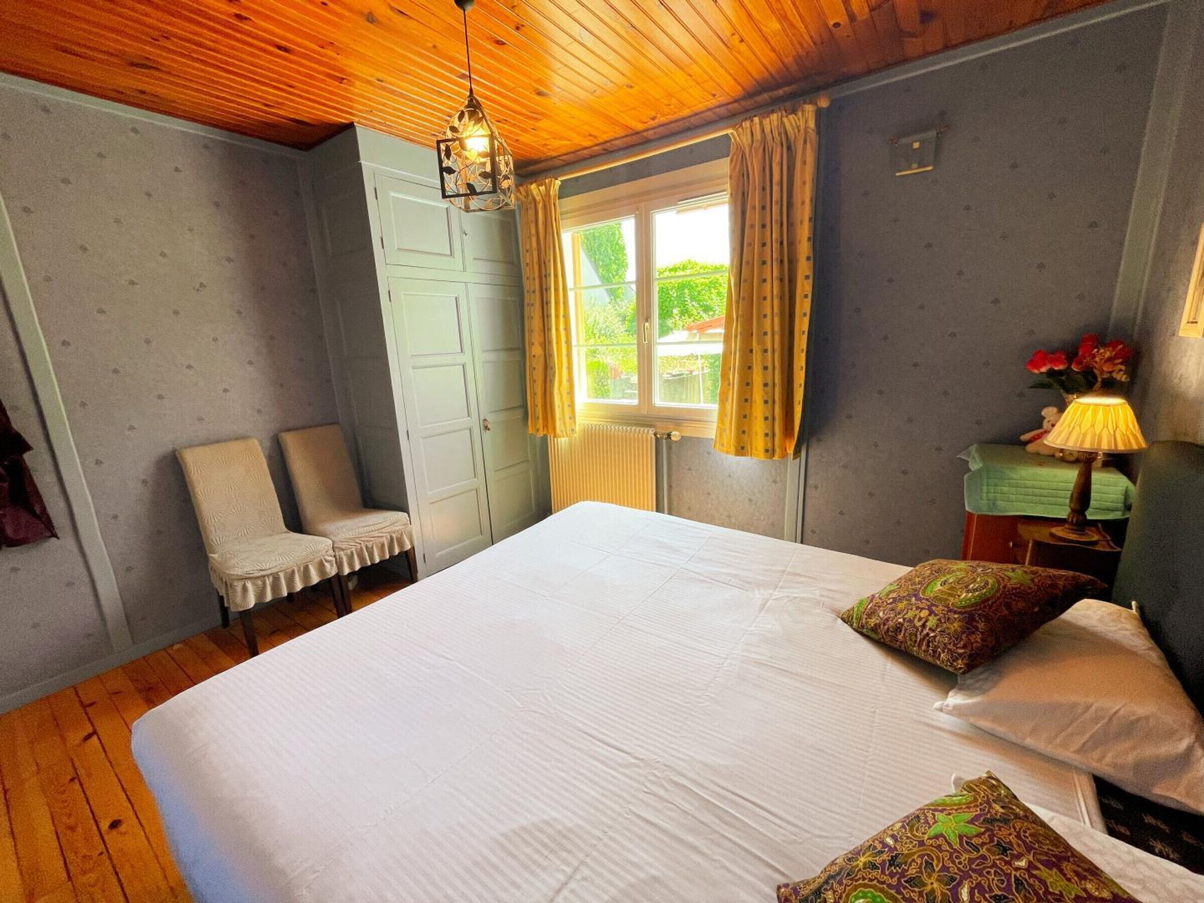 Un dormitorio con techo de madera, una cama con ropa de cama blanca, una silla, un ventanal con cortinas y una mesita de noche con lámpara.
