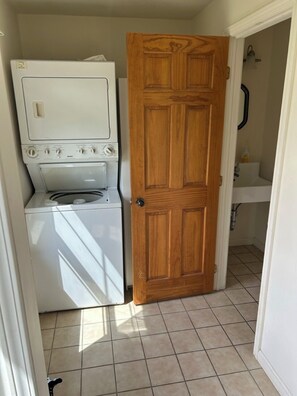 Un espace buanderie équipé d’un lave-linge et d’un sèche-linge, une porte en bois et une salle de bain avec un lavabo et un miroir.