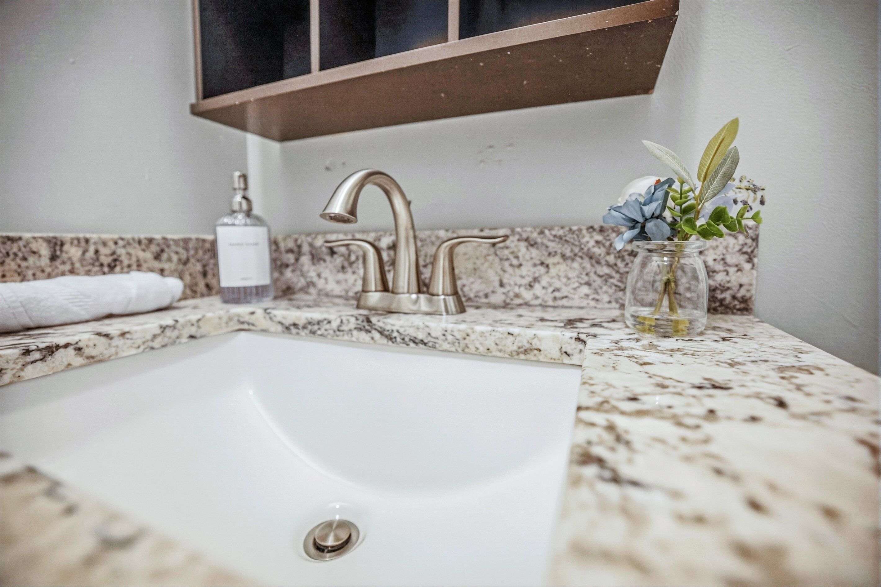 A bathroom with a white sink, a single faucet, a marble countertop, a glass vase with flowers, and a bottle of hand soap.