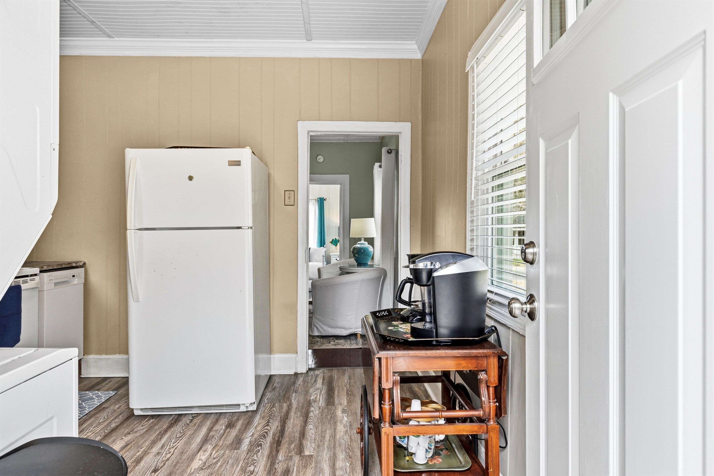 A compact kitchen with a white refrigerator, a coffee maker on a wooden stand, and a door leading to a bathroom.
