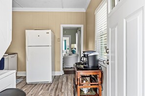 A compact kitchen with a white refrigerator, a coffee maker on a wooden stand, and a door leading to a bathroom. - Haymont Hideaway- A timeless stay in the Heart of Haymount! (Fayetteville)