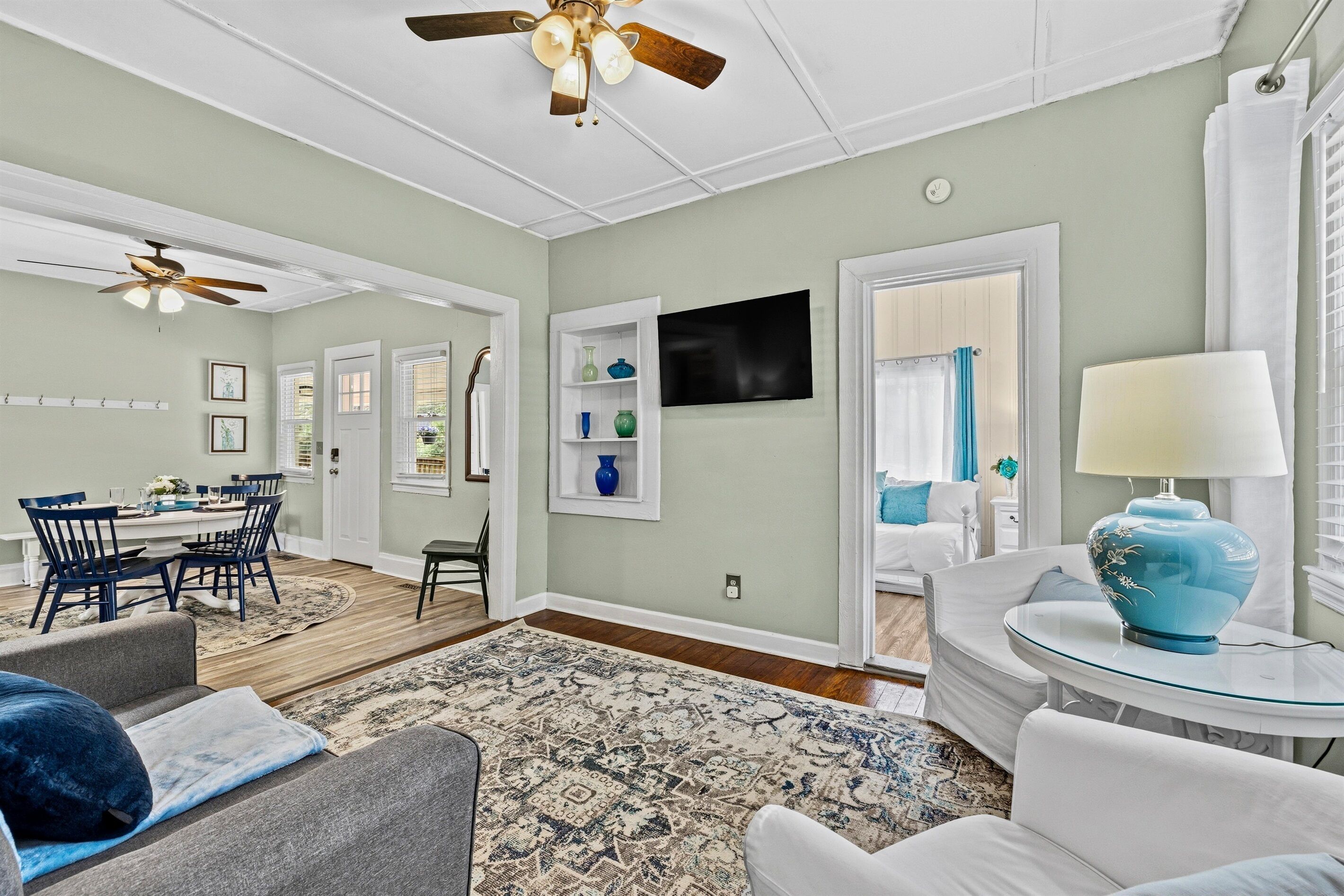 A spacious living room with a dining area, a television mounted on the wall, and a ceiling fan.