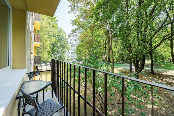 A balcony with a table and chairs, overlooking a park with trees and a building in the background. - RentPlanet - Apartament Joliot-Curie (Warszawa)