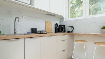 Una cuina moderna amb armaris blancs, tauleig de fusta, microones integrat, cafetera i una finestra amb vista a arbres.