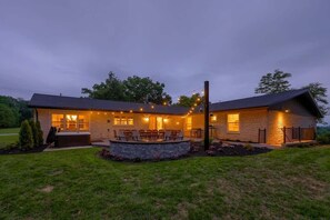 A two-story house with a stone foundation, a covered patio with seating, and a well-lit exterior.