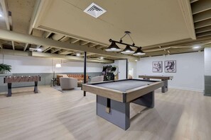 A pool table in a room with a high ceiling, recessed lighting, and a foosball table.