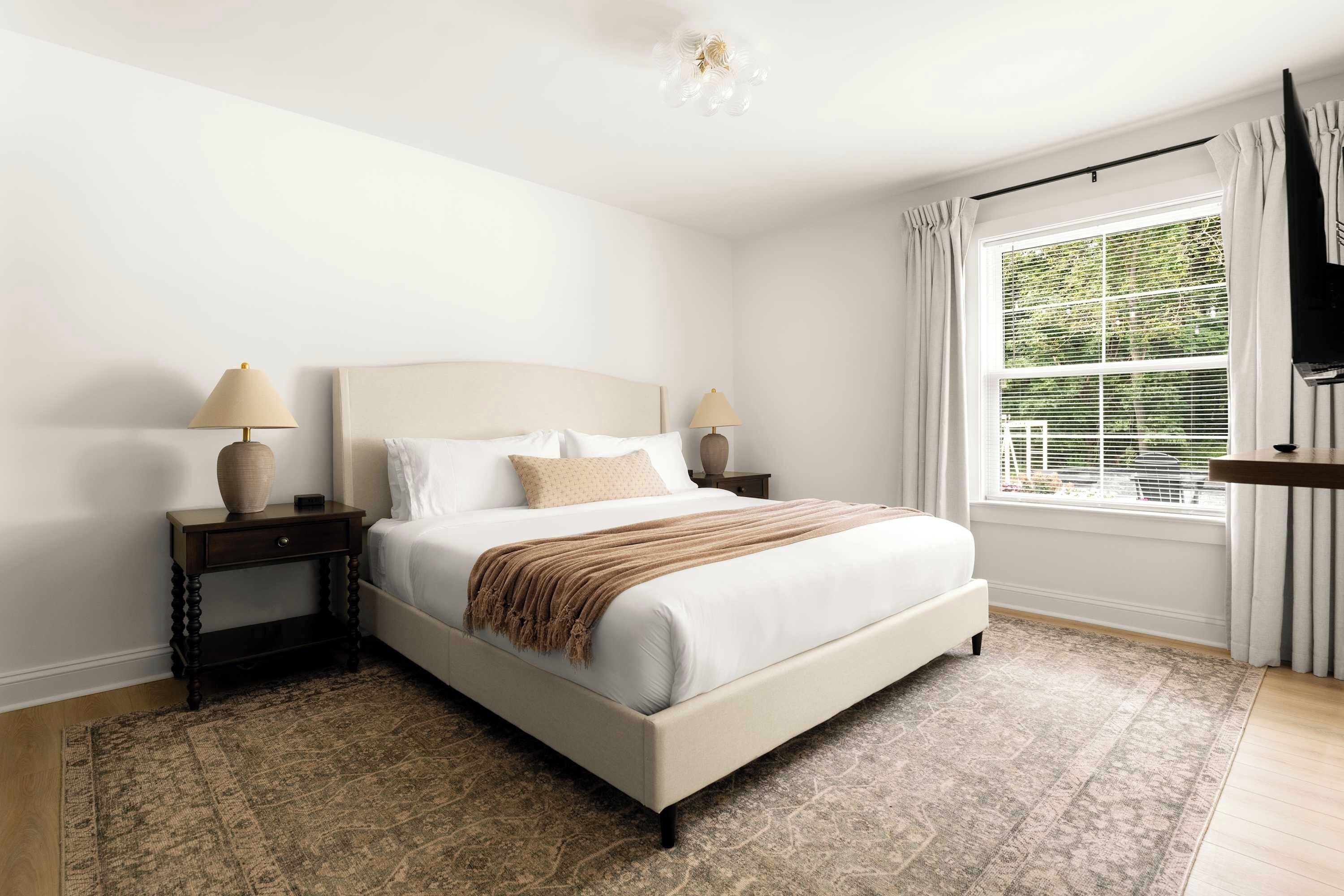 Une chambre à coucher comprenant un lit, des tables de chevet, un tapis, une fenêtre avec des stores et un luminaire au plafond.
