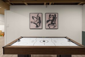 A pool table with a wooden frame and a white playing surface, flanked by two abstract black paintings on a white wall.