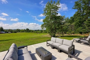 An outdoor seating area with a sofa and chairs, a fire pit, and a view of a grassy field and trees.