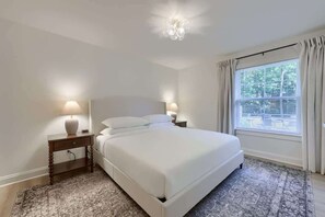 A neatly made bed in a room with a large window, two bedside lamps, and a patterned rug.