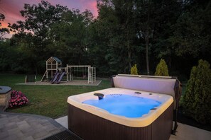 A hot tub in a backyard with a swing set and a playhouse in the background.