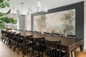 A dining room with a large wooden table surrounded by chairs, a painting on the wall, and a window with curtains.