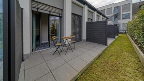 A modern residential building with a patio area featuring a table and chairs, a sliding glass door, and a fenced garden.