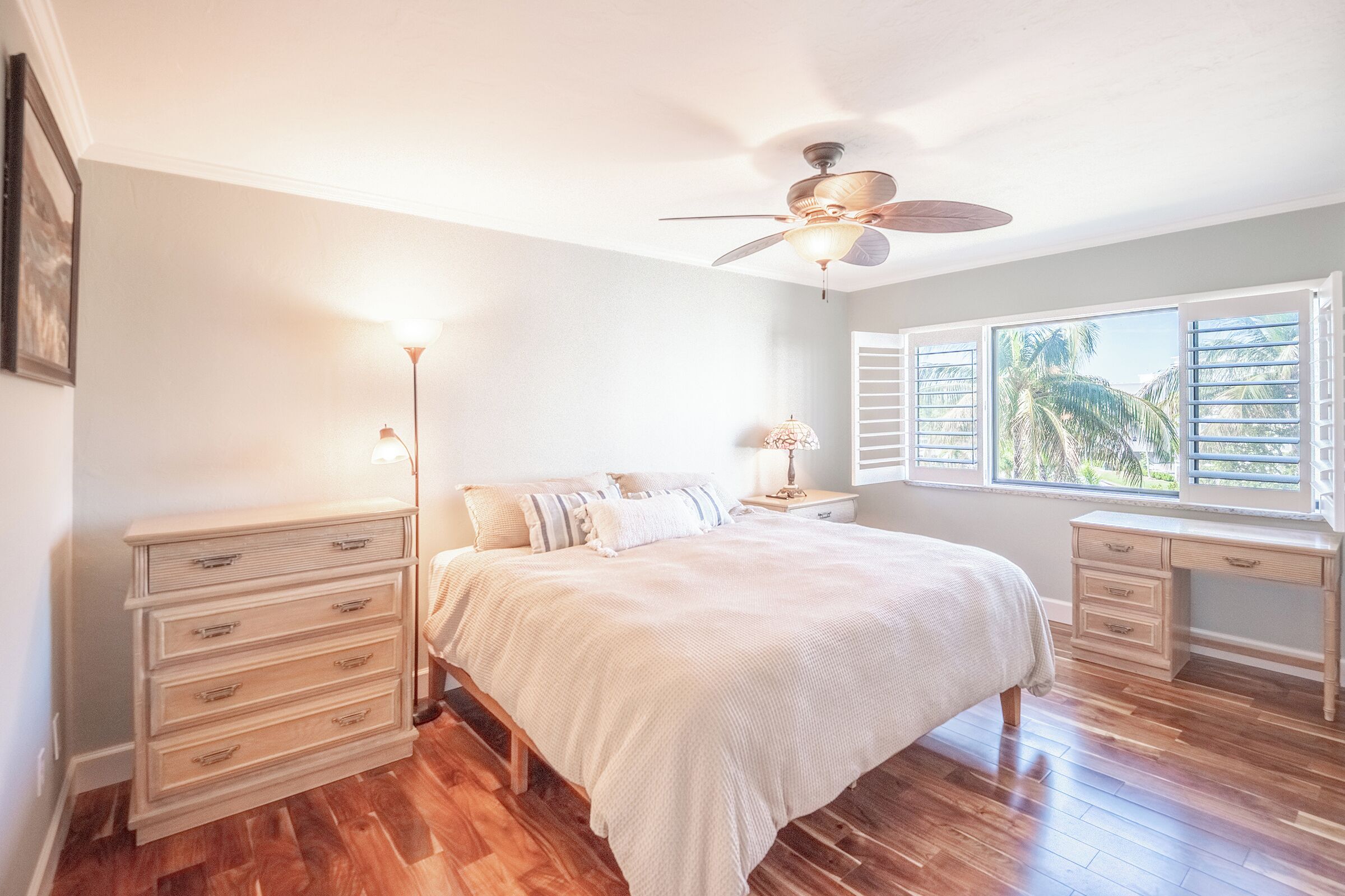 Quarto com cama, mesas de cabeceira, escrivaninha, ventoinha no teto e janela com persianas.