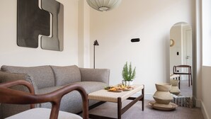 Ein modernes Wohnzimmer mit einer Couch, einem Holzstuhl, einem Couchtisch mit Dekoration, einer Stehlampe und einem Spiegel, der den Raum reflektiert.