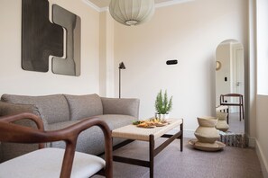 Ein modernes Wohnzimmer mit einer Couch, einem Holzstuhl, einem Couchtisch mit Dekoration, einer Stehlampe und einem Spiegel, der den Raum reflektiert.